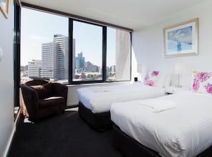 Quarto de hotel com duas camas, uma cadeira, uma janela ampla com vista para a cidade e um quadro na parede.