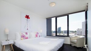 A modern hotel room with a large bed, a chair, a nightstand, a lamp, and a window with a city view. - Exclusive Stays - Yarra Condos (Southbank)