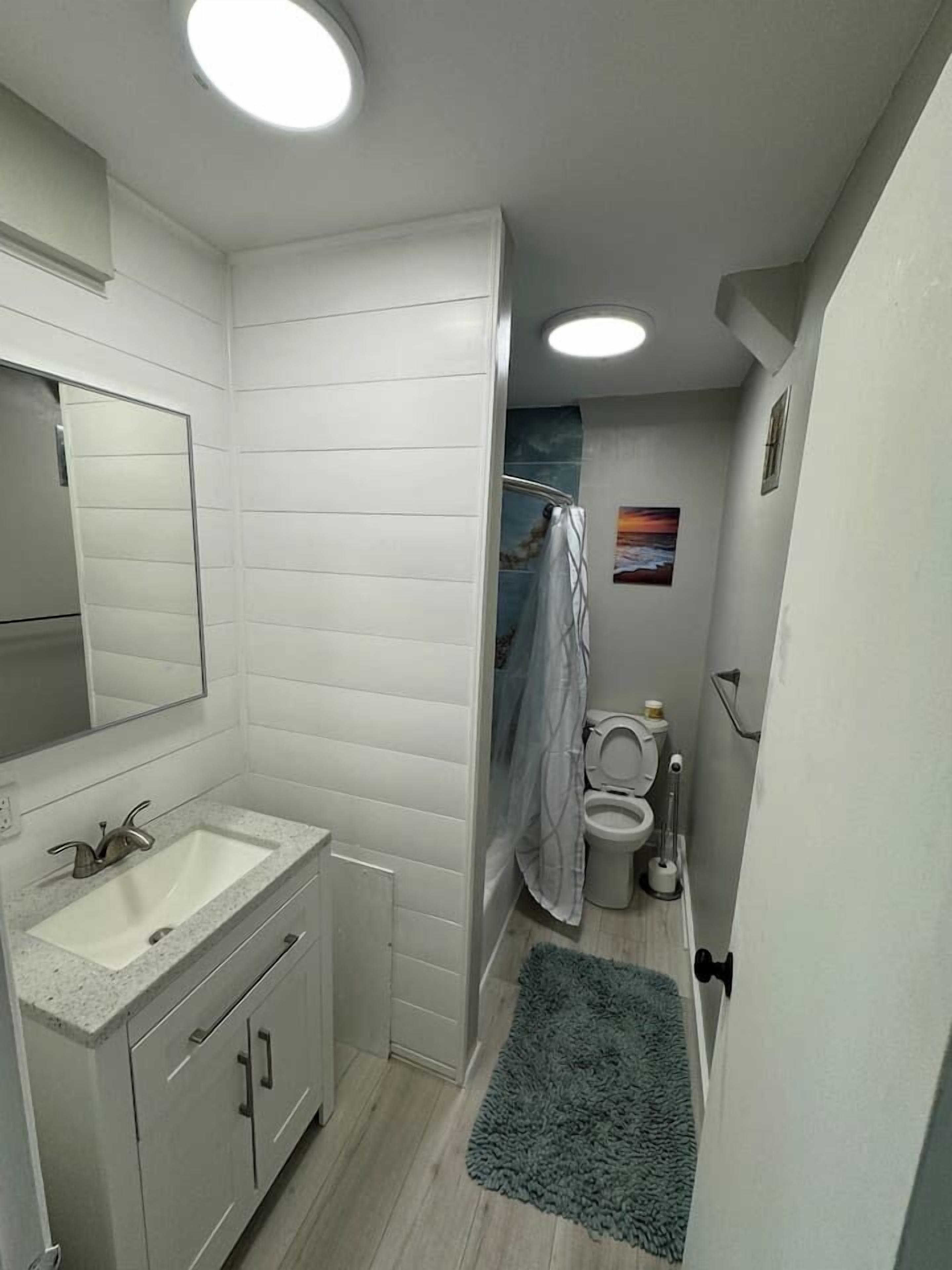 A bathroom with a sink, mirror, toilet, shower curtain, and a small rug.
