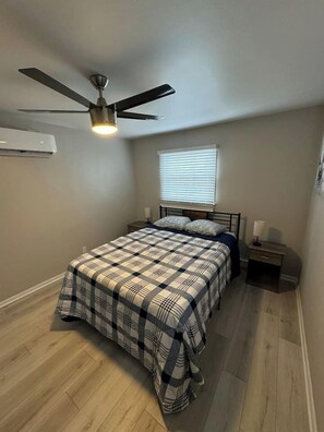 Een slaapkamer met een bed, een plafondventilator, een raam met zonwering, een nachtkastje en een airconditioning.