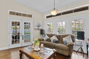 Ein großzügiges Wohnzimmer mit einem braunen Sofa, einem Couchtisch mit Blumenvase und einem großen Fenster mit Blick auf Grünflächen.