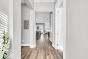 A hallway with white walls, a wooden floor, and a door leading to a living area. - Panama City Beach, FL | 6BR | 4.5BA | Sleeps 18 | Private Pool | Dog-Friendly (Panama City Beach)