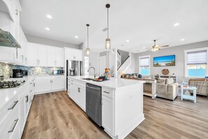 A modern kitchen with white cabinetry, a central island, and a dining area with a sofa and table. - Panama City Beach, FL | 6BR | 4.5BA | Sleeps 18 | Private Pool | Dog-Friendly (Panama City Beach)