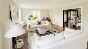 Un salón amplio con un sofá blanco, una mesa de centro y una lámpara. Hay una puerta que da a otra habitación y una escalera.
