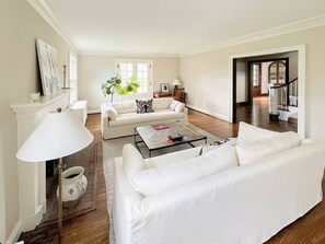 Ein geräumiges Wohnzimmer mit einem weißen Sofa, einem Couchtisch und einer Lampe. Eine Tür führt in einen anderen Raum, und eine Treppe ist ebenfalls zu sehen.