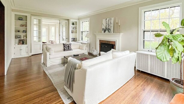 Un salón amplio con suelos de madera, muebles blancos, chimenea y ventanas grandes.