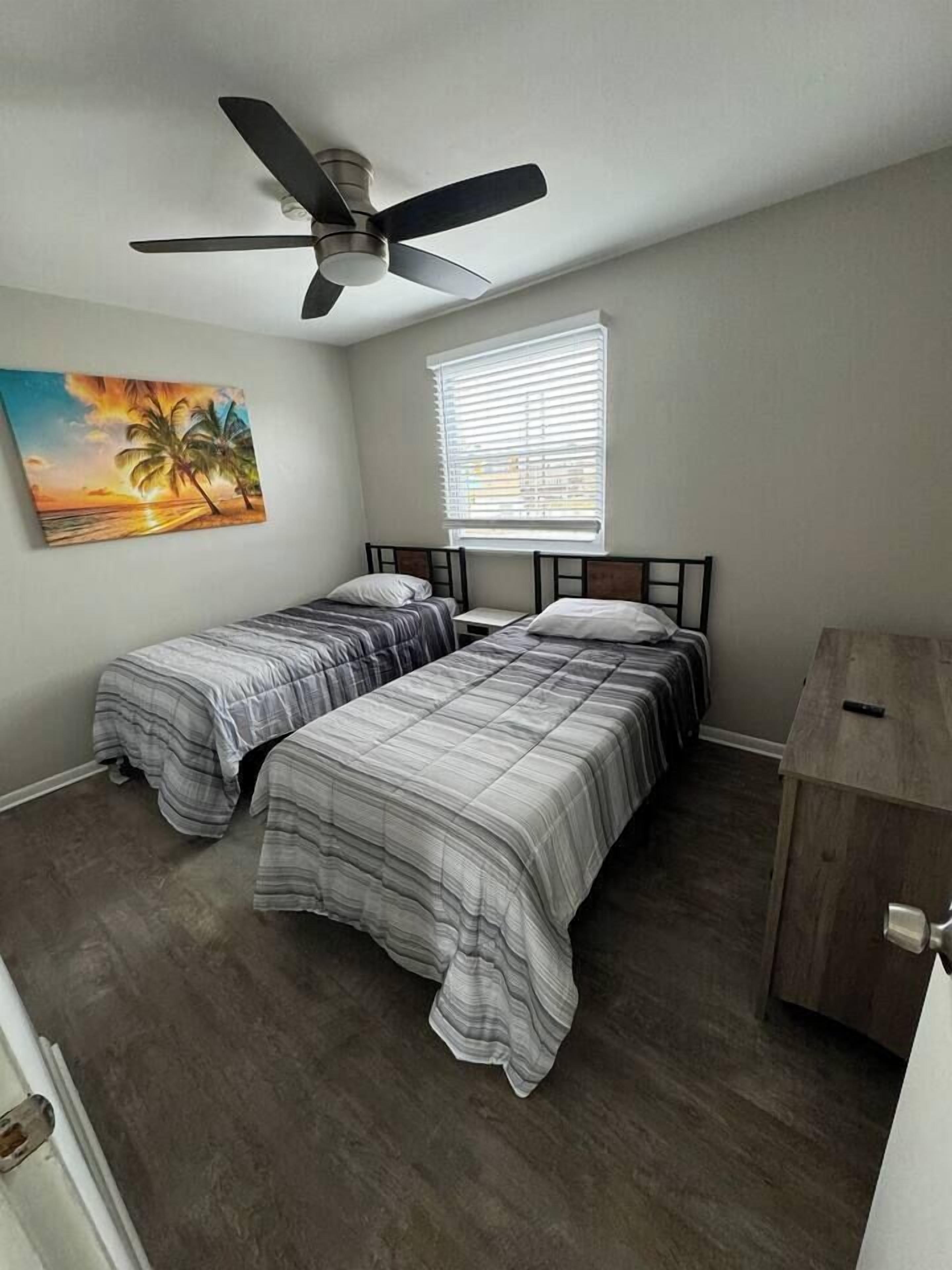 Une chambre avec deux lits, un ventilateur de plafond, une fenêtre avec des stores, une table de chevet en bois et un tableau au mur.