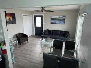 Una sala de estar con un sofá oscuro, una mesa de centro de vidrio, un sillón pequeño y un ventilador de techo.