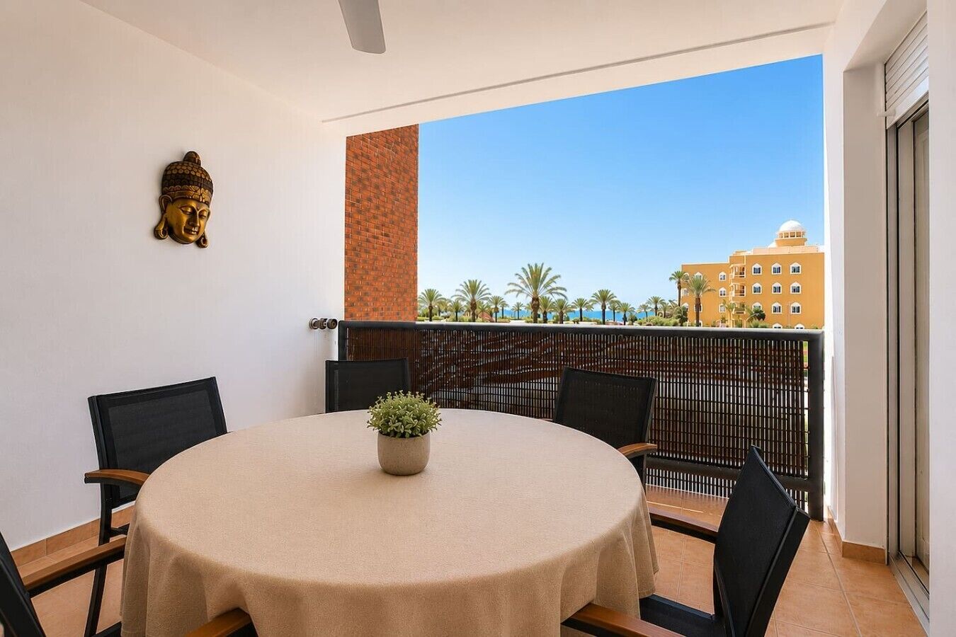 A balcony with a round table, chairs, a Buddha statue, and a view of buildings and palm trees.