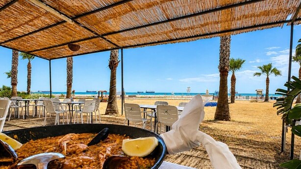 Prato de paella de frutos do mar sobre uma mesa, com vista para a praia e palmeiras.