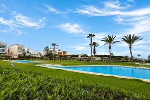 Ein Schwimmbecken mit blauem Himmel, Palmen und modernen Gebäuden im Hintergrund.