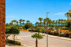 A parking area with palm trees, a clear sky, and a distant view of a building with a pool.