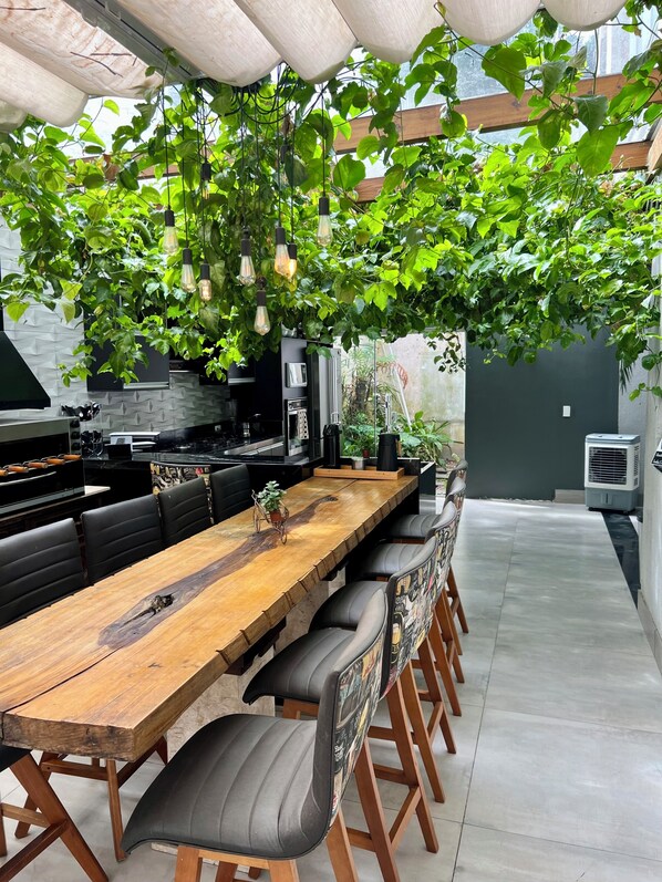 Un área de comedor al aire libre con una larga mesa de madera, sillas con cojines negros, lámparas colgantes y una estación de parrilla.
