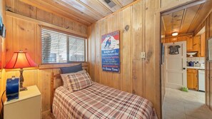 A wooden room with a bed, a lamp, a poster, and a door leading to a kitchen. - Regan Beach Retreat Cabin | CYH0809 (South Lake Tahoe)