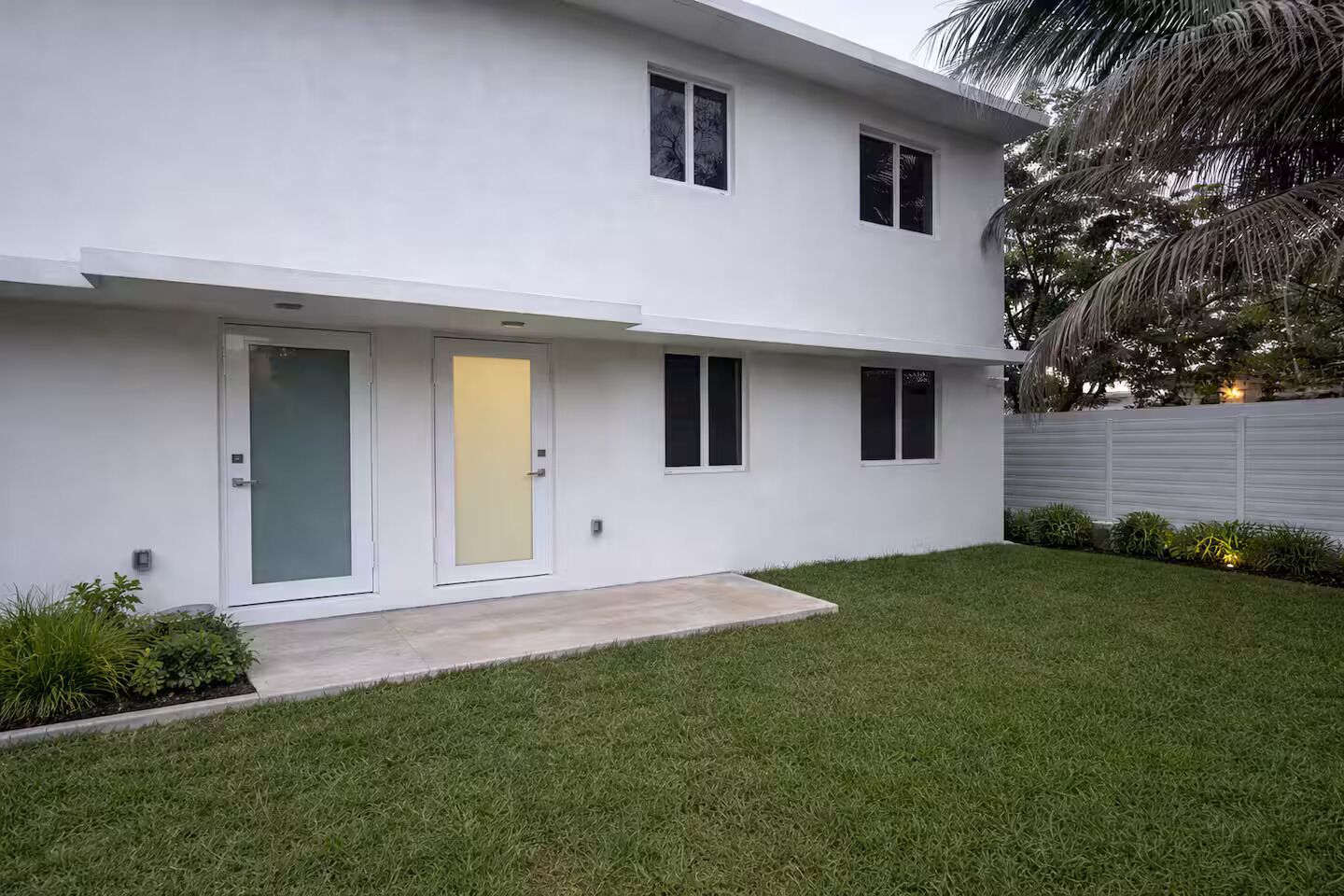 Una casa moderna de dos pisos con techo plano, puerta principal y pequeño jardín delantero con césped y plantas.