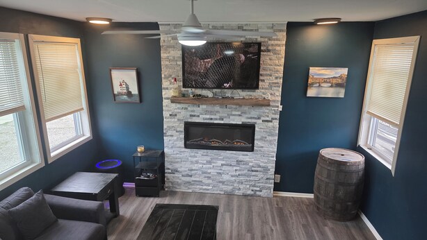 A living room with a stone fireplace, a ceiling fan, and a large window with blinds. - Lake Erie Fishing - Sleeps 8 · 3 Bd - 1.5 Bth - Full Kitchen - Patio - Out Elec. (Lorain)