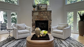 A living room with a stone fireplace, two sofas, a coffee table, and large windows with a view of trees.