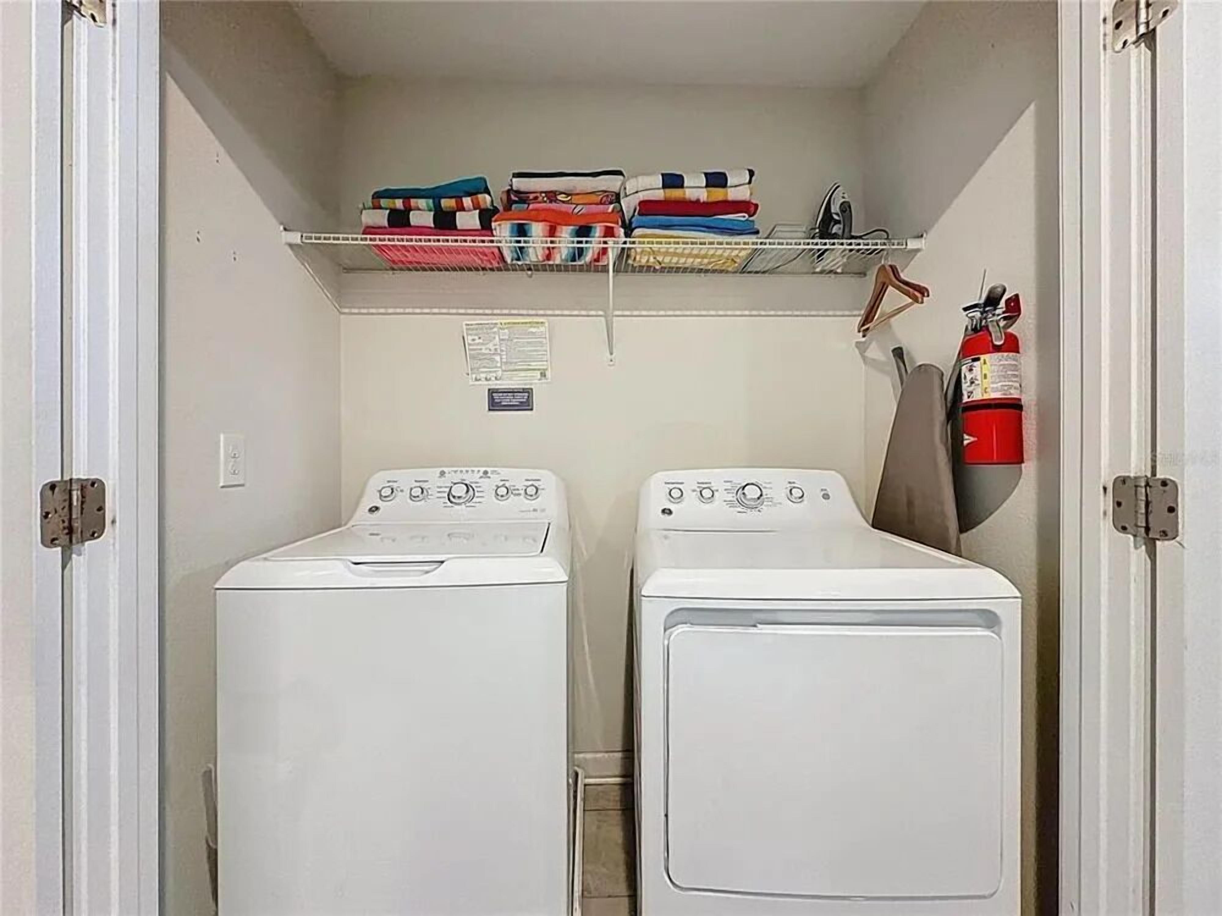 Een wasserette met twee witte voorladers, wasmachines en drogers, een metalen plank met opgevouwen dekens, een brandblusser en een bericht op de muur.