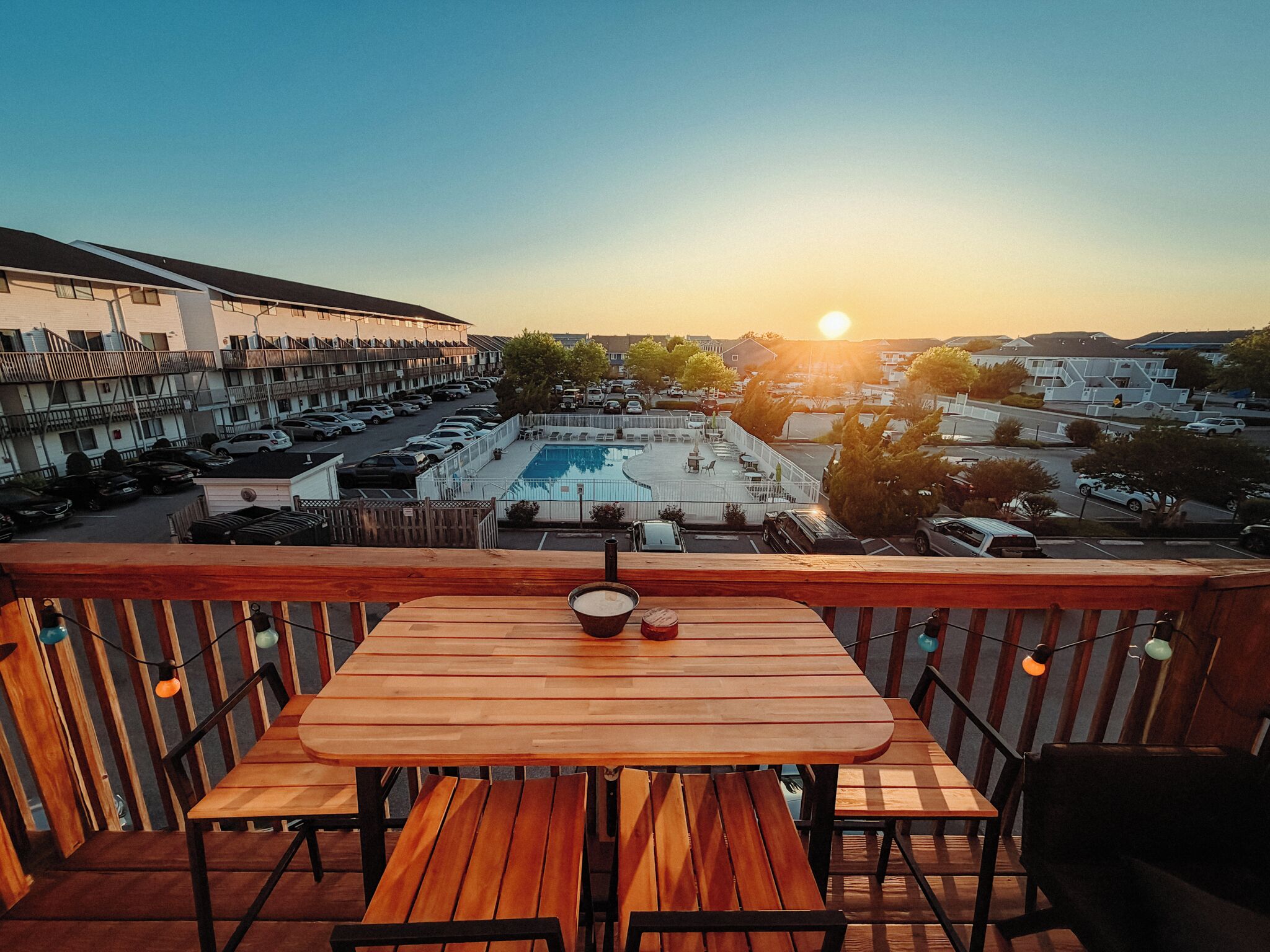 Een houten tafel en stoelen op een balkon met uitzicht op een zwembad en een parkeerplaats.