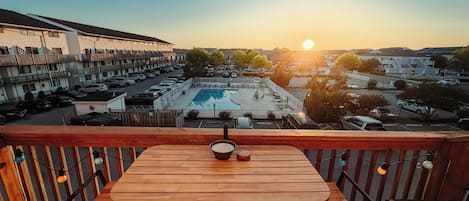 A wooden table and chairs on a balcony overlooking a swimming pool and a parking lot.