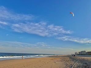 Ein Strand mit blauem Himmel, eine Person am Wasser und Gebäude in der Ferne.