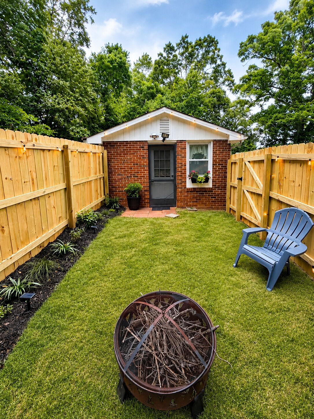 A backyard with a small brick house, a wooden fence, a blue chair, and a fire pit.