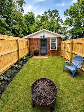A backyard with a small brick house, a wooden fence, a blue chair, and a fire pit.