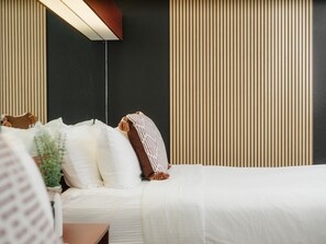 Una cama bien hecha con ropa blanca, una mesita de noche con lámpara y una pared con listones de madera verticales.