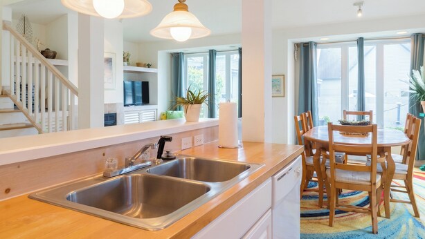A modern kitchen with stainless steel sinks, a wooden dining table, and a staircase with white railings. - Charming 1-bedroom Downtown Escape in Saugatuck, perfect for solo travelers. (Saugatuck)