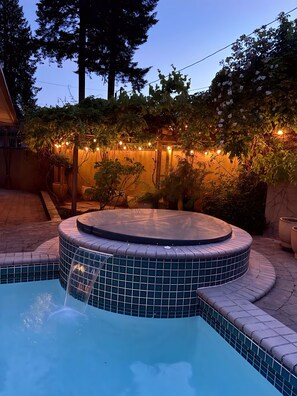 Un jacuzzi con borde de azulejos, rodeado por un patio de ladrillo y luces colgantes de fondo.