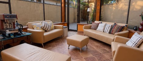 A sunlit room with a glass ceiling, beige sofas, a coffee station, and a view of trees.