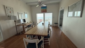 Een eetgedeelte met een lange houten tafel, witte stoelen, een plafondventilator en uitzicht door de ramen.