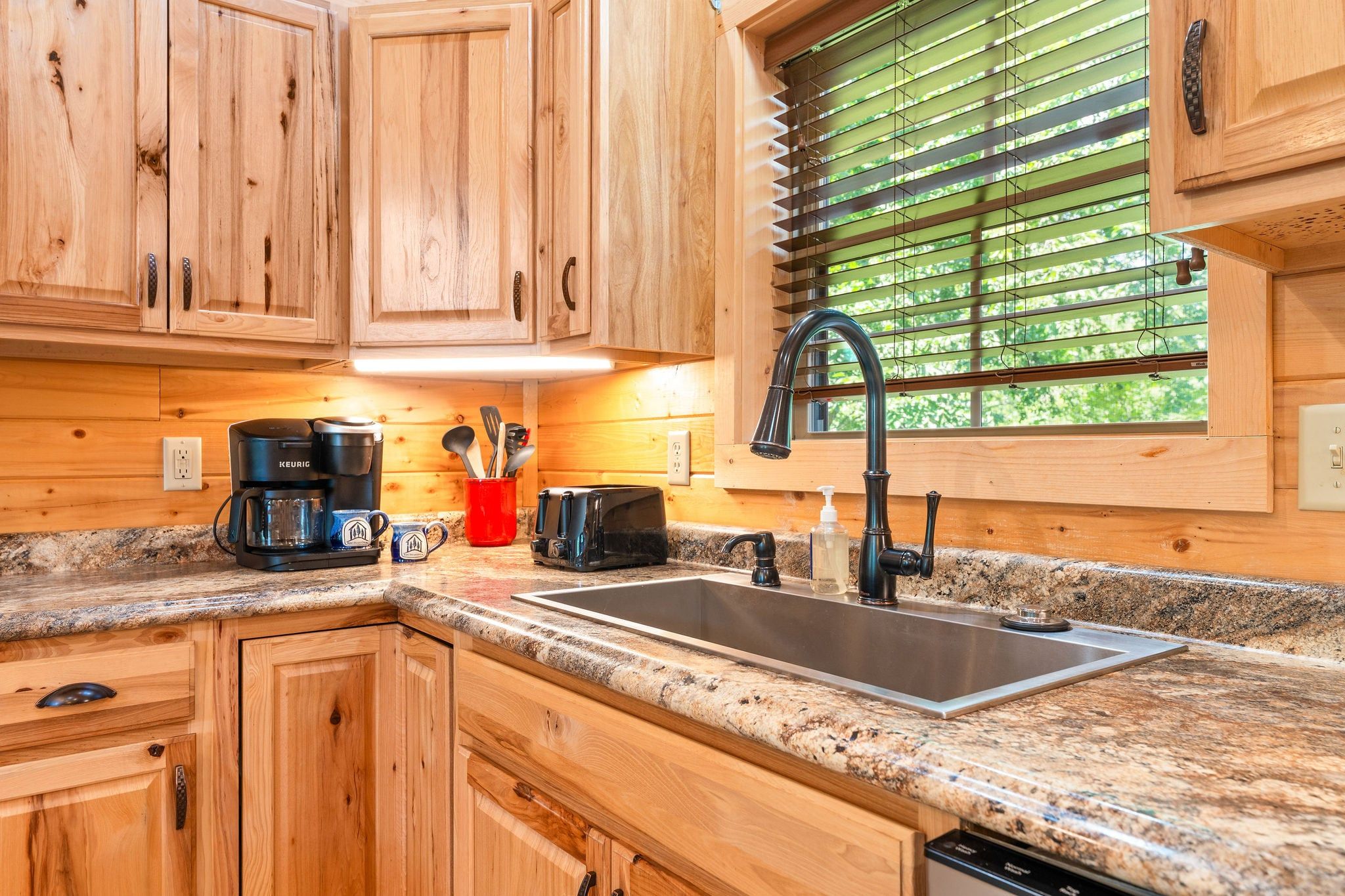 A kitchen with wooden cabinets, a granite countertop, a coffee maker, a toaster, and a window with blinds.