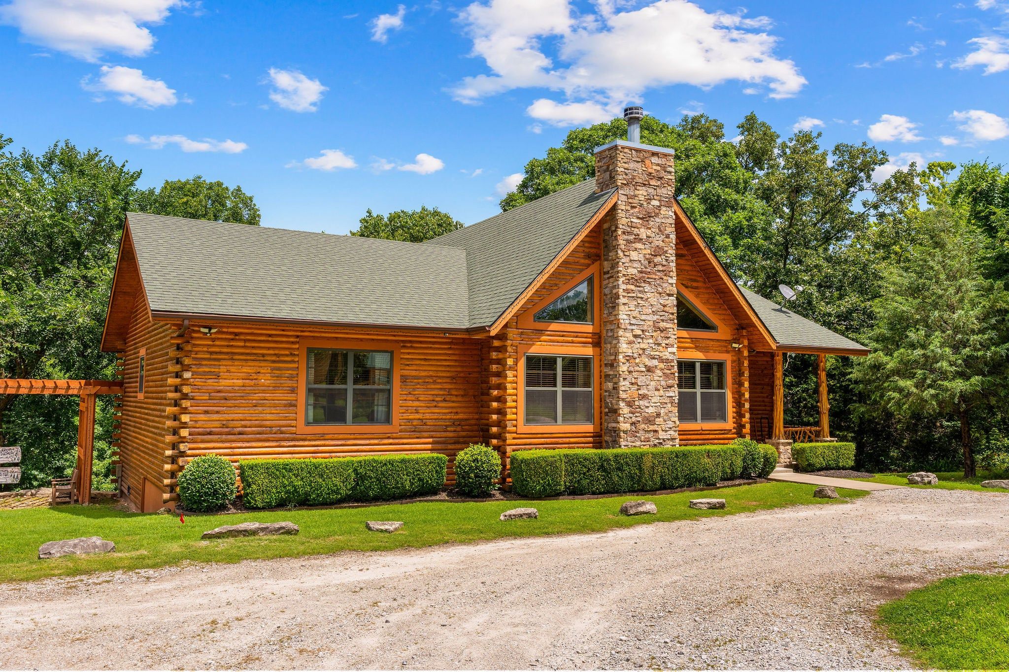 A log cabin with a stone chimney, large windows, and a gravel driveway.