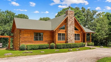 A log cabin with a stone chimney, large windows, and a gravel driveway.