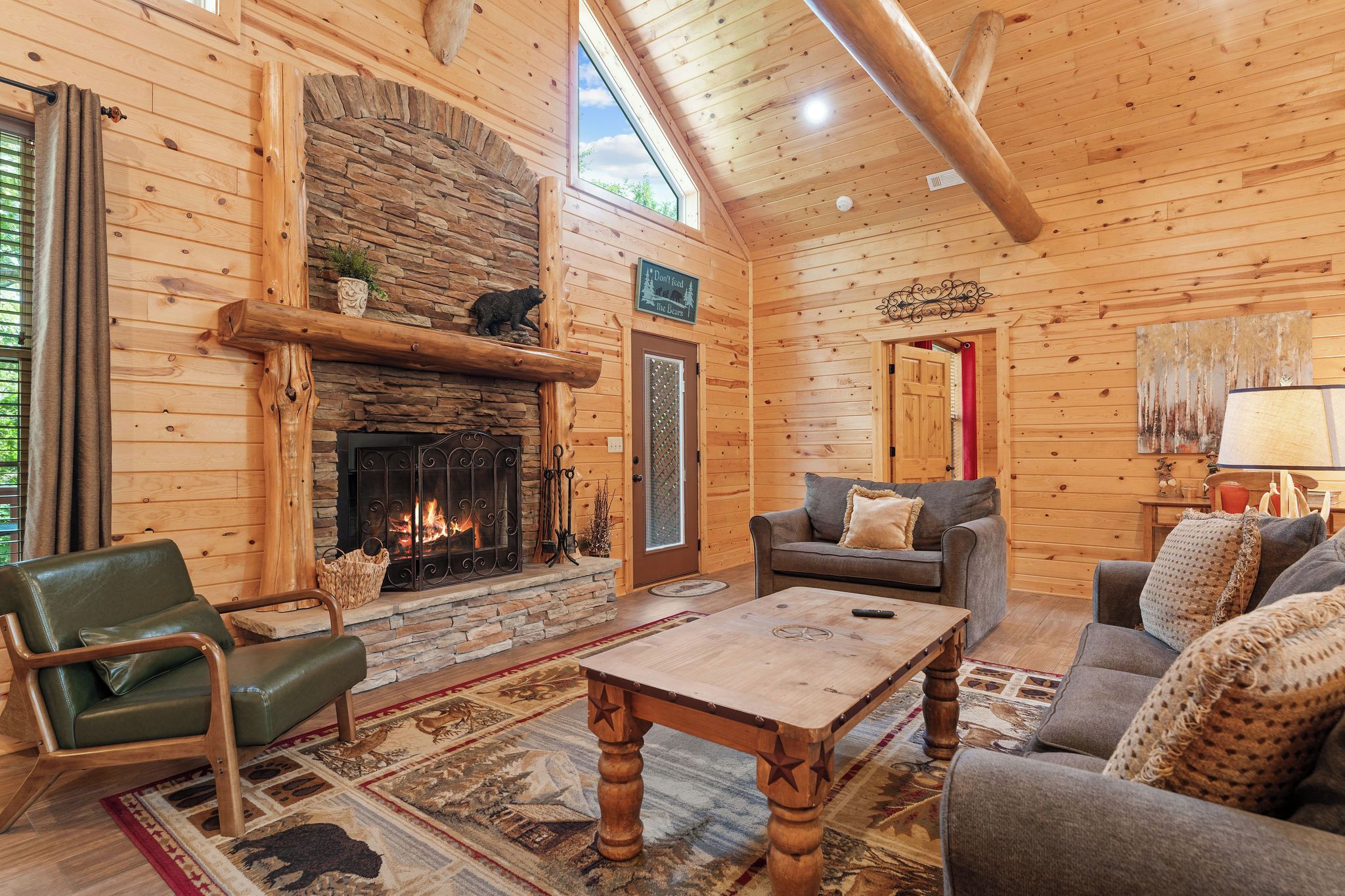 A log cabin interior with a stone fireplace, wooden furniture, and a large window.
