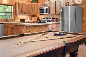 A pool table with pool cues and balls, wooden cabinets, a microwave, a stove, and a refrigerator in a kitchen area. - Secluded Ozark Log Cabin w/ Private Fire Pit, Hot Tub & Pool Table (Ridgedale)