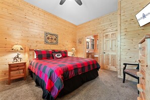 A wooden-paneled room with a bed, a nightstand, a lamp, a bench, and a door. - Secluded Ozark Log Cabin w/ Private Fire Pit, Hot Tub & Pool Table (Ridgedale)