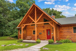 A log cabin with a red door, a porch with a bench and chairs, and a stone pathway leading to the entrance. - Secluded Ozark Log Cabin w/ Private Fire Pit, Hot Tub & Pool Table (Ridgedale)