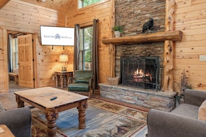 A cozy cabin interior with a stone fireplace, wooden walls, a television mounted on the wall, and a comfortable seating area with a table and chairs. - Secluded Ozark Log Cabin w/ Private Fire Pit, Hot Tub & Pool Table (Ridgedale)