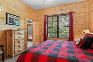 Ein Zimmer in einer Holzhütte mit einem Bett, einem Schminktisch, einem Fenster mit Vorhängen und einem gerahmten Bild an der Wand.