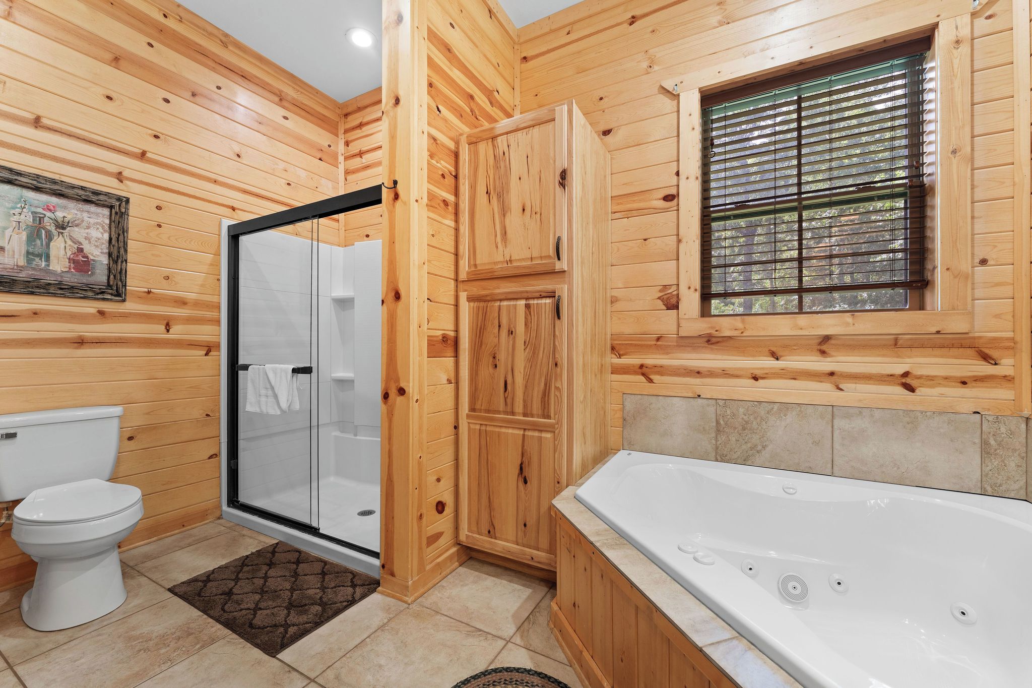 A wooden cabin bathroom with a glass shower, a white toilet, a bathtub, and a window with blinds.