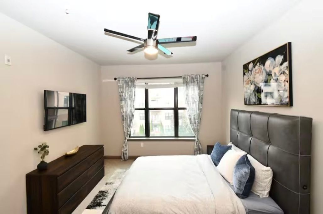 Une chambre à coucher avec un lit, un ventilateur de plafond, une fenêtre avec des rideaux, un téléviseur à écran plat, une commode en bois foncé et un tableau accroché au mur.