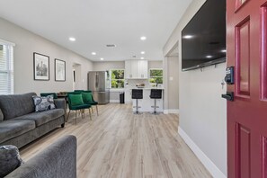 A modern living room with a grey sofa, green chairs, and a kitchen with white cabinets and a red door.