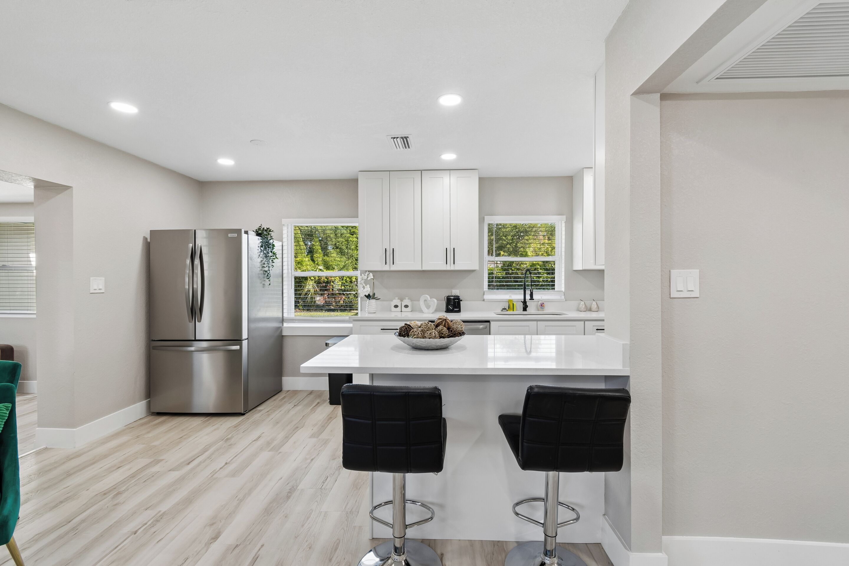 A modern kitchen with a stainless steel refrigerator, white cabinets, a central island with two black bar stools, and a window with a view of greenery.