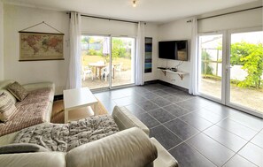 A living room with a sofa, a coffee table, a flat-screen TV, and a sliding glass door leading to an outdoor patio. - Charmante maison en Vendée (Moutiers-Les-Mauxfaits)