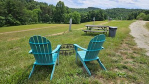 Two Adirondack chairs, a picnic table, and a trash can in a grassy area with a forested hill in the background.