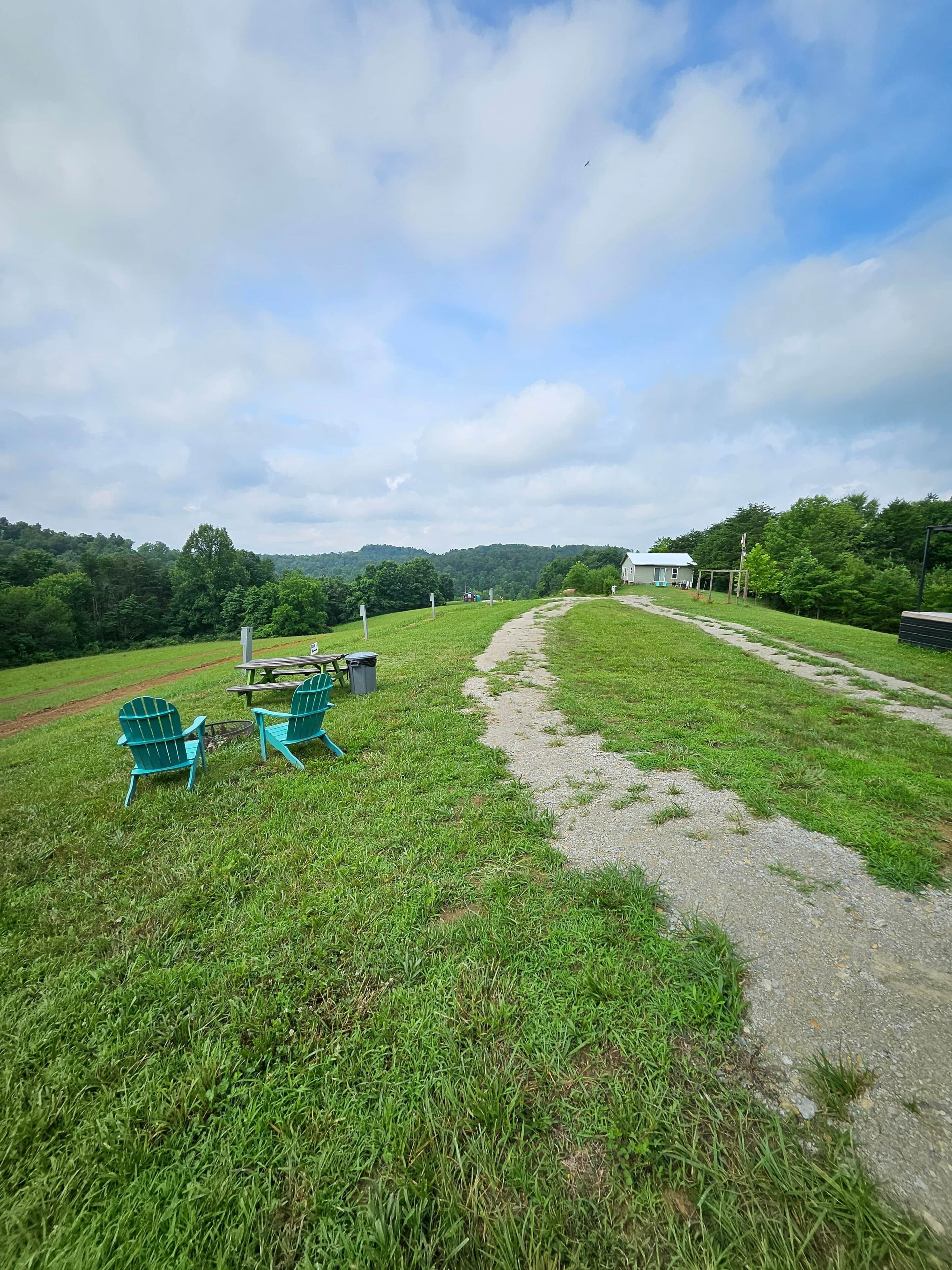 A grassy field with a dirt path, two green chairs, and a small table with a trash bin.