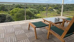 A balcony with a wooden floor, two wooden chairs with green cushions, a small table with a white pitcher and a cup, and a view of a green landscape. - Get away at Rivoli Bay Farm Stay. Beach access close by. Sleeps 6 (Beachport)