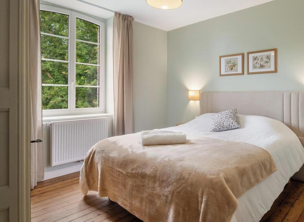 Un dormitorio con un ventanal grande, una cama con un edredón color crema, un cabecero y dos marcos de cuadros colgados en la pared.